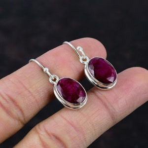 以下が含まれることがあります： 楕円形のルビーの宝石が付いたシルバーのイヤリングのペア。イヤリングは、フック留め具でシンプルなデザインです。