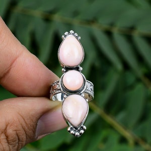 May include: A silver ring featuring three light pink gemstones. The top and bottom stones are teardrop-shaped, while the center stone is round. The ring has intricate detailing around the gemstones and on the band.