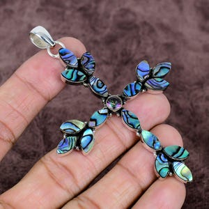 May include: A silver-toned cross pendant with iridescent abalone shell accents. The cross features a central gemstone and leaf-shaped shell pieces in shades of blue, green, and purple. The pendant has a bail for a chain.