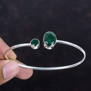 May include: A silver bangle bracelet with two oval-shaped emerald green gemstones. The larger gemstone is on the right side of the bracelet and the smaller gemstone is on the left side.