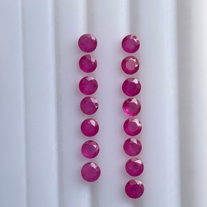 May include: A collection of sixteen round, faceted, deep red rubies. The gemstones are arranged in two vertical columns of eight, set against a white, ridged surface. The rubies exhibit a vibrant, polished appearance.