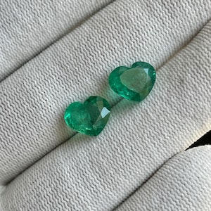 May include: Two heart-shaped emerald gemstones. The gems are a vibrant green colour and have a faceted cut, reflecting light. They are displayed on a white, textured surface.