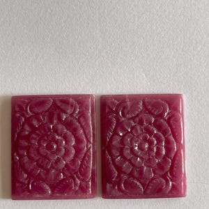Puede incluir: Dos cabujones rectangulares de piedra preciosa de color rosa intenso con un motivo floral tallado. Cada pieza muestra una flor central en flor rodeada de hojas estilizadas, con una superficie lisa y pulida y bordes definidos en tono magenta.