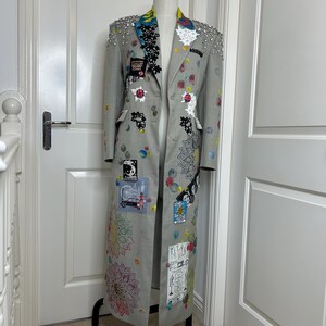 Può includere: Un cappotto lungo grigio con toppe colorate, schizzi di vernice e abbellimenti. Il cappotto presenta un colletto a revers con fodera fantasia ed è adornato con strass sulle spalle. È esposto su un manichino nero.