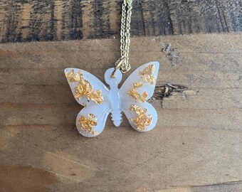 Puede incluir: Un collar con colgante de mariposa con una cadena dorada. El colgante de mariposa es blanco con detalles de pan de oro. El collar se muestra sobre una superficie de madera.