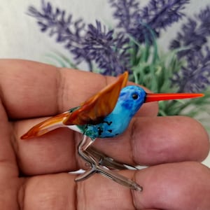 May include: A small, glass hummingbird figurine with a blue body, orange wings, and a long, red beak. The bird has detailed features and sits on clear metal legs. The background includes purple flowers.