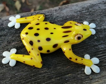 Könnte beinhalten: Eine gelbe Froschfigur mit schwarzen Punkten und weißen Füßen. Der Frosch liegt auf dem Rücken mit ausgestreckten Beinen.