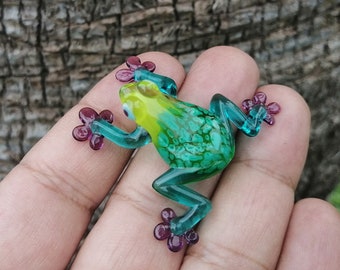 Könnte beinhalten: Eine kleine Glasfroschfigur mit einem lebendigen Farbschema. Der Frosch hat einen gelben Kopf, einen grünen Körper mit einem strukturierten Muster und türkisfarbene Beine und Füße. Die Füße haben lila, blütenartige Details. Die Figur wird in einer Hand gehalten.