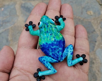 Könnte beinhalten: Eine kleine blaue und grüne Froschfigur mit schwarzen Augen und Füßen. Der Frosch ist mit einem gefleckten Muster bemalt.