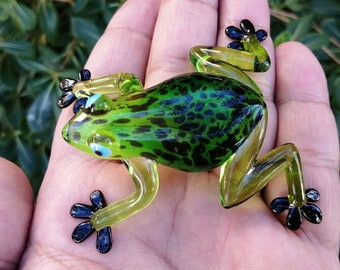 Könnte beinhalten: Eine grüne Glasfröschchenfigur mit schwarzen Flecken und schwarzen Füßen. Der Frosch sitzt auf einer Hand.