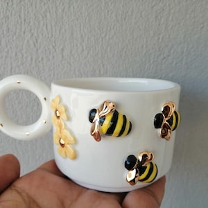 Op de afbeelding: Witte keramische mok met een handvat en gouden accenten. De mok heeft drie zwarte en gele bijen en gele bloemen.