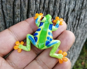 Könnte beinhalten: Eine kleine, grüne, blaue und gelbe Froschfigur mit orangenen Füßen. Der Frosch sitzt auf einer Hand.