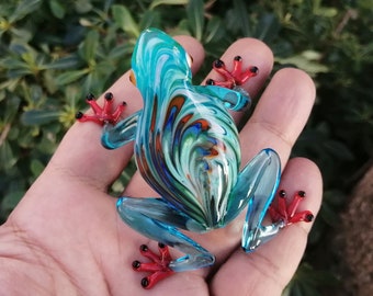 May include: A glass frog figurine with a blue and green swirl pattern and red accents. The frog is sitting on a hand.