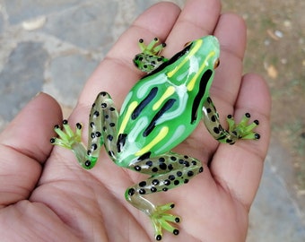 Könnte beinhalten: Eine grüne Glasfröschchenfigur mit schwarzen und gelben Streifen und schwarzen Punkten. Der Frosch sitzt auf einer Hand.