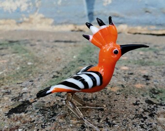 May include: A glass figurine of a hoopoe bird with an orange body, black and white stripes, and a long, thin beak. The bird is standing on a rough, brown surface.