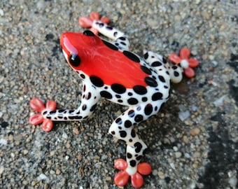 May include: A decorative frog figurine with a vibrant red head and back, contrasted by a white body speckled with black spots. The frog's feet are a coral-like color, and it has black eyes. The figurine is set against a textured, gray surface.