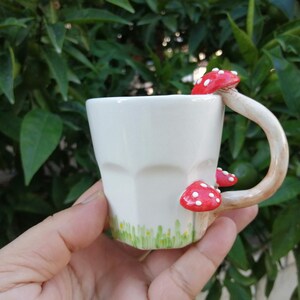 May include: White ceramic mug with a mushroom-shaped handle. The handle is painted to look like a mushroom with a red cap and white spots. The mug has a green and white design around the bottom.