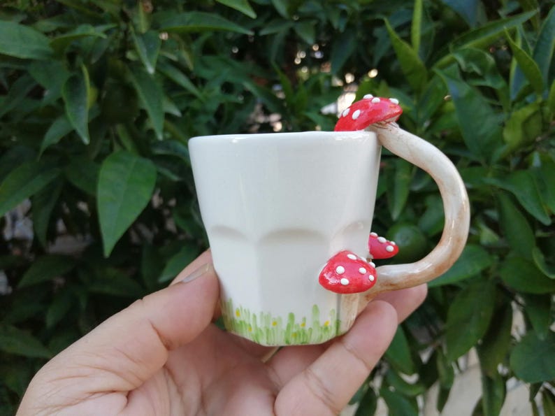 May include: White ceramic mug with a mushroom-shaped handle. The handle is painted to look like a mushroom with a red cap and white spots. The mug has a green and white design around the bottom.