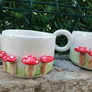 May include: Two white ceramic mugs with red and white mushroom designs. The mugs have handles and are decorated with a green and white background.