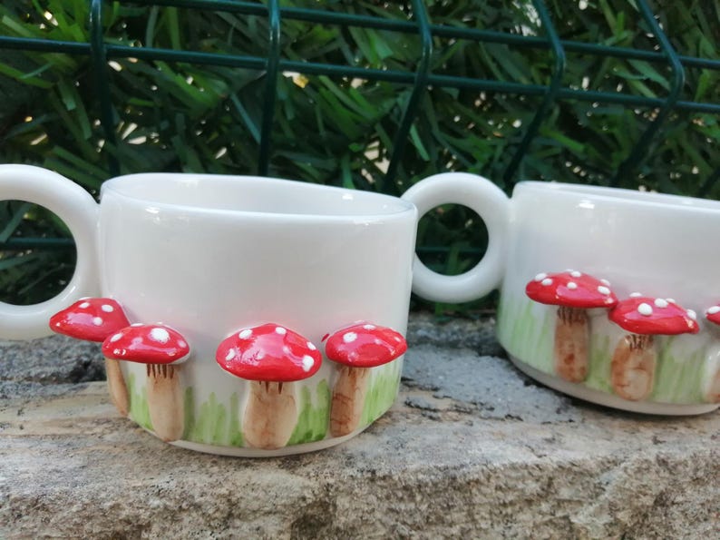 May include: Two white ceramic mugs with red and white mushroom designs. The mugs have handles and are decorated with a green and white background.