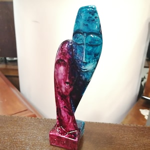 May include: Abstract sculpture of two faces, one blue and one pink, intertwined and joined at the forehead. The sculpture is made of a smooth, glossy material.