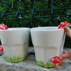May include: Two white ceramic mugs with mushroom handles. The mugs have a green and yellow design around the bottom. The mushrooms are red with white spots.