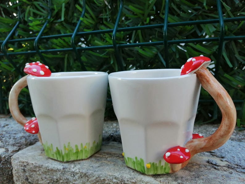 May include: Two white ceramic mugs with mushroom handles. The mugs have a green and yellow design around the bottom. The mushrooms are red with white spots.