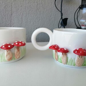 May include: Two white ceramic mugs with red and white mushroom designs. The mugs have a handle and are decorated with a row of mushrooms with red caps and white spots.