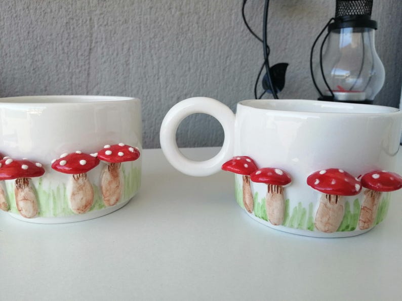 May include: Two white ceramic mugs with red and white mushroom designs. The mugs have a handle and are decorated with a row of mushrooms with red caps and white spots.