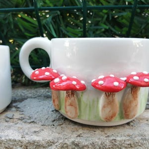 May include: White ceramic mug with red and white mushroom decorations. The mushrooms have white spots and are arranged around the mug.