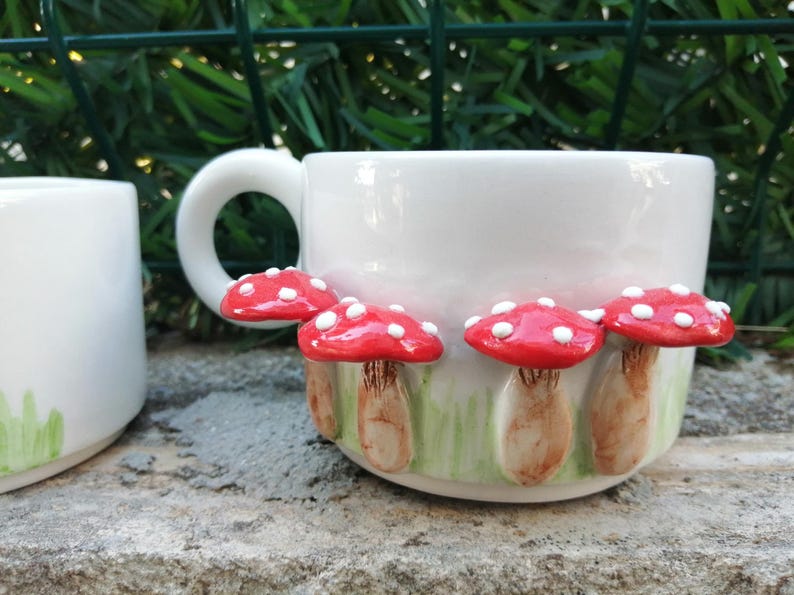May include: White ceramic mug with red and white mushroom decorations. The mushrooms have white spots and are arranged around the mug.