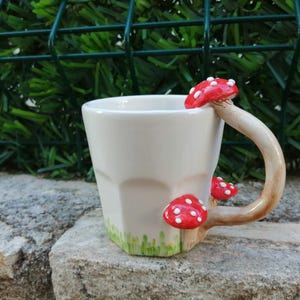 May include: White ceramic mug with a handle shaped like a mushroom. The mug has three red and white mushrooms attached to the handle. The mug is sitting on a rock.