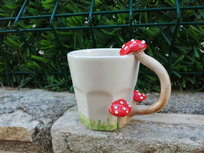 May include: White ceramic mug with a handle shaped like a mushroom. The mug has three red and white mushrooms attached to the handle. The mug is sitting on a rock.