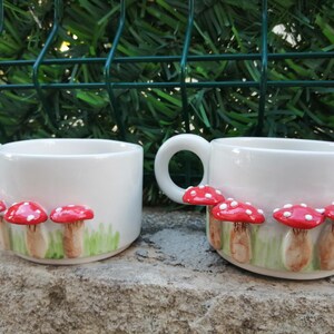 May include: Two white ceramic mugs with red and white mushroom designs. The mushrooms are 3D and have a green grass background.