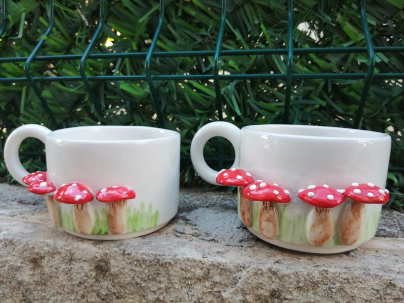 May include: Two white ceramic mugs with red and white mushroom designs. The mushrooms are 3D and have a green grass background.