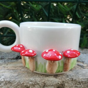 May include: White ceramic mug with a handle and four red and white mushrooms attached to the side.