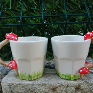 May include: Two white ceramic mugs with mushroom handles. The mugs have a red and white mushroom on the handle and a green and yellow grass design at the bottom.