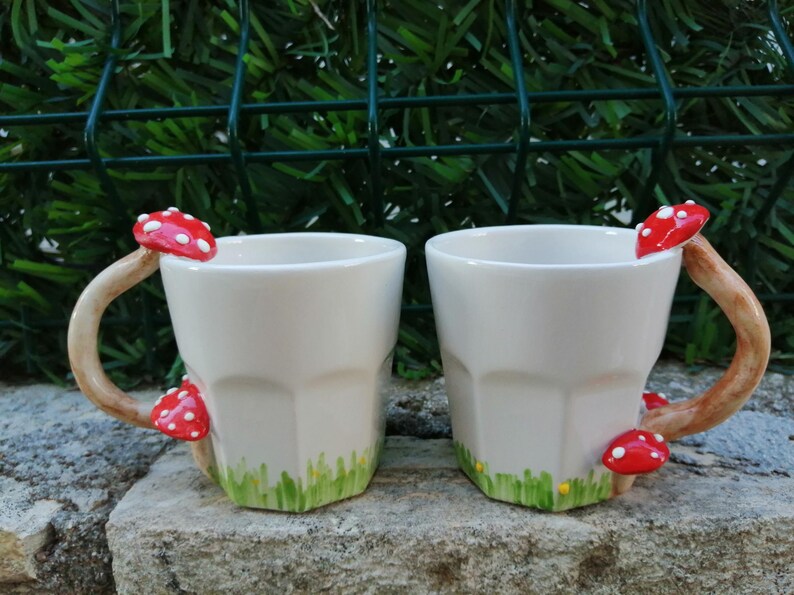 May include: Two white ceramic mugs with mushroom handles. The mugs have a red and white mushroom on the handle and a green and yellow grass design at the bottom.