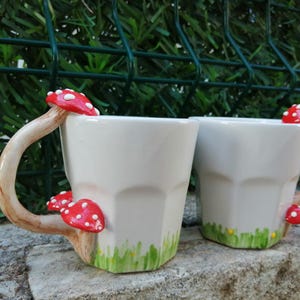 May include: Two white ceramic mugs with mushroom handles. The mugs have a green and yellow painted grass design at the bottom. The mushroom handles are red with white spots.