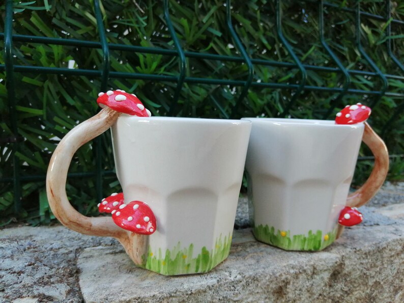 May include: Two white ceramic mugs with mushroom handles. The mugs have a green and yellow painted grass design at the bottom. The mushroom handles are red with white spots.