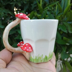 May include: White ceramic mug with a mushroom-shaped handle and red and white mushroom accents. The mug has a green and white design on the bottom.