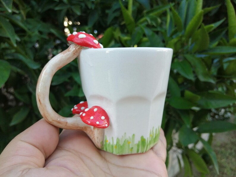 May include: White ceramic mug with a mushroom-shaped handle and red and white mushroom accents. The mug has a green and white design on the bottom.