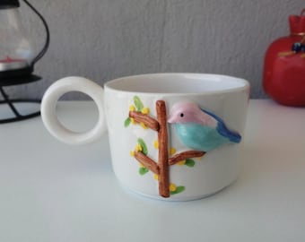 May include: White ceramic mug with a large, round handle. The mug features a 3D sculpted bird with blue and pink feathers perched on a brown branch with yellow berries and green leaves. The mug is a decorative item.