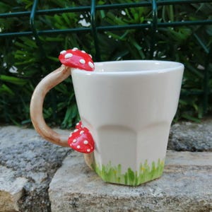 May include: White ceramic mug with a mushroom-shaped handle and red and white mushroom accents. The mug has a green and white grass design around the bottom.