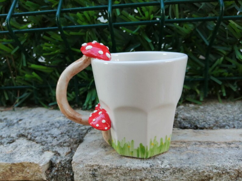 May include: White ceramic mug with a mushroom-shaped handle and red and white mushroom accents. The mug has a green and white grass design around the bottom.