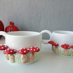 May include: Two white ceramic mugs with red and white mushroom designs. The mugs have a rounded shape and a handle on the side.