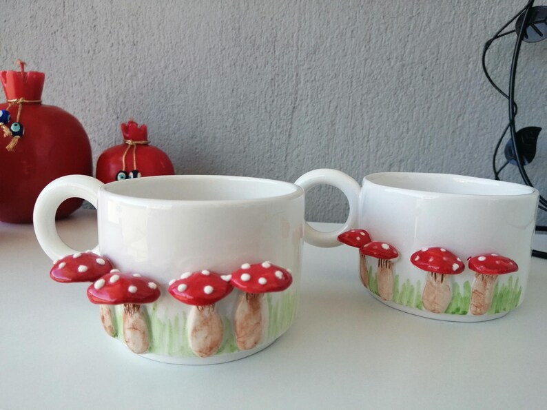 May include: Two white ceramic mugs with red and white mushroom designs. The mugs have a rounded shape and a handle on the side.