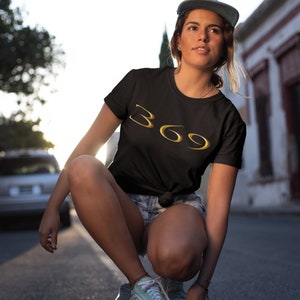 May include: A black t-shirt with a gold 369 graphic design. The person wearing the shirt is squatting down and looking to the side.