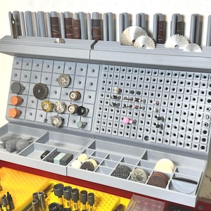 Puede incluir: Un organizador de herramientas gris, montado en la pared, con múltiples compartimentos y un diseño de panel perforado. Contiene varios accesorios para herramientas rotativas, incluyendo tambores de lijado, hojas de sierra y brocas de esmerilado. El organizador está diseñado para mantener las herramientas.