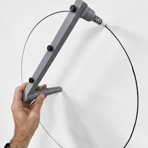 May include: A gray drawing compass tool with black knobs and an attached EXPO marker is used to draw a black circle on a white surface. The tool is held by a person's hand.